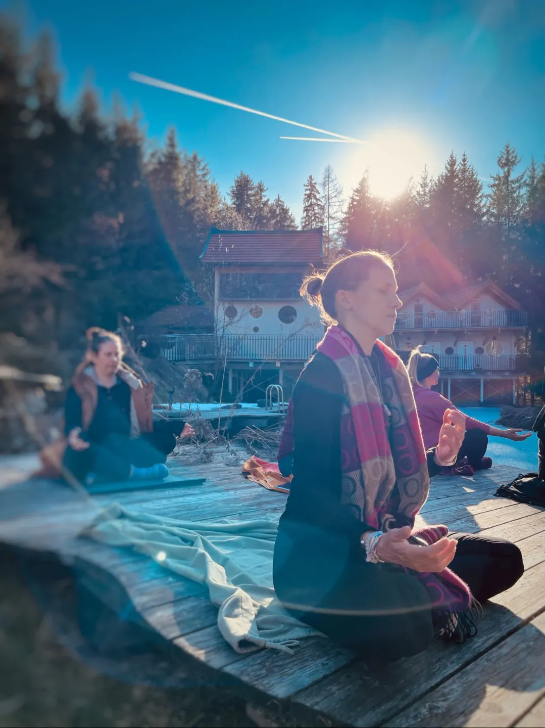 Stefanie Jahn meditierend am Steg zusammen mit anderen Frauen, die Sonne und ein Gebäude im Hintergrund